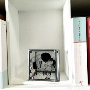May include: A white bookshelf with books on it. The books are in different languages and have different titles. There is a glass cube on the shelf with a black and white image of a couple inside.