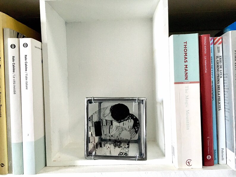 May include: A white bookshelf with books on it. The books are in different languages and have different titles. There is a glass cube on the shelf with a black and white image of a couple inside.