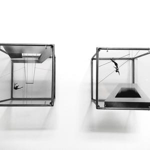 May include: Two clear glass boxes with metal frames, each containing a figure suspended by wires. The figure in the left box is hanging from a swing, while the figure in the right box is suspended in mid-air.