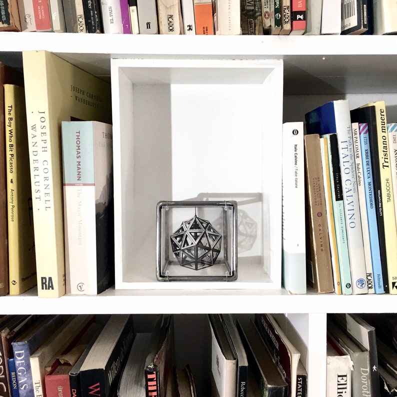 May include: A white bookshelf with books on each shelf. The top shelf has a variety of books with different titles and authors. The middle shelf has a clear glass cube with a silver geometric shape inside. The bottom shelf has a variety of books with different titles and authors.