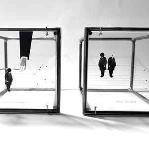 Könnte beinhalten: Zwei Glas-Ausstellungsboxen mit schwarzen Metallrahmen. Eine Box zeigt eine kleine Figur unter großen Händen, mit dem Text "Infectious Thoughts". Die andere Box zeigt zwei Figuren, mit dem Text "Mirror Thoughts".