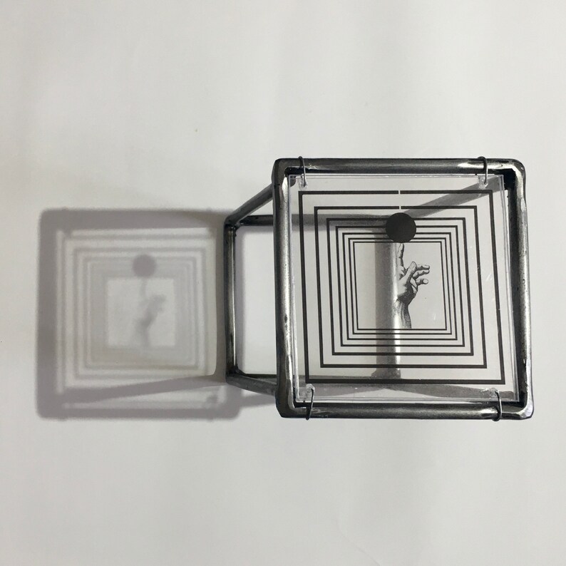 May include: A three-dimensional cube sculpture constructed from dark metal frames. The top face features a clear square panel printed with concentric black squares, a solid black circle, and a detailed illustration of a hand reaching upward.