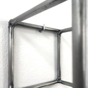 May include: A square-shaped wall-mounted shelf constructed from dark grey metal tubing. The frame features visible welded joints at each corner and includes a small, silver-colored metal hook attached to the top horizontal bar for hanging items.
