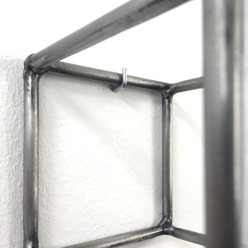 May include: A square-shaped wall-mounted shelf constructed from dark grey metal tubing. The frame features visible welded joints at each corner and includes a small, silver-colored metal hook attached to the top horizontal bar for hanging items.