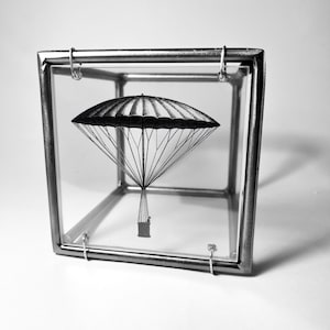 Puede incluir: Una ilustración en blanco y negro de un globo aerostático dentro de un cubo de vidrio. El globo tiene una cesta y está suspendido por cuerdas.
