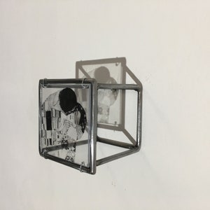 May include: A black and white image of a couple kissing is framed in a silver metal cube. The cube is mounted on a white wall.