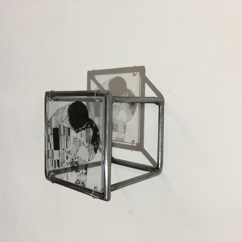 May include: A black and white image of a couple kissing is framed in a silver metal cube. The cube is mounted on a white wall.