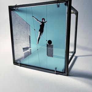 May include: A square glass panel suspended in a dark metal frame featuring a graphic illustration of a swimmer in a black swimsuit and red cap diving into a light blue pool with a gray concrete wall, a black sphere, and thin black geometric lines.