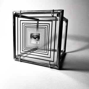 Op de afbeelding: Een driedimensionaal glazen kubussculptuur met een reeks in elkaar grijpende zwarte vierkante contouren op het voorpaneel. In het midden van deze concentrische vierkanten bevindt zich een gedetailleerde zwart-wit grafische illustratie van een menselijk oog.