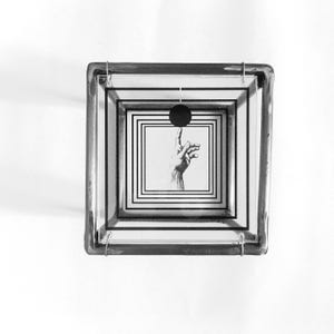 May include: A square glass box with a metal frame featuring a layered design of black concentric squares. Inside, a black circle is positioned above an illustration of a hand with an extended index finger, creating a visual depth effect.