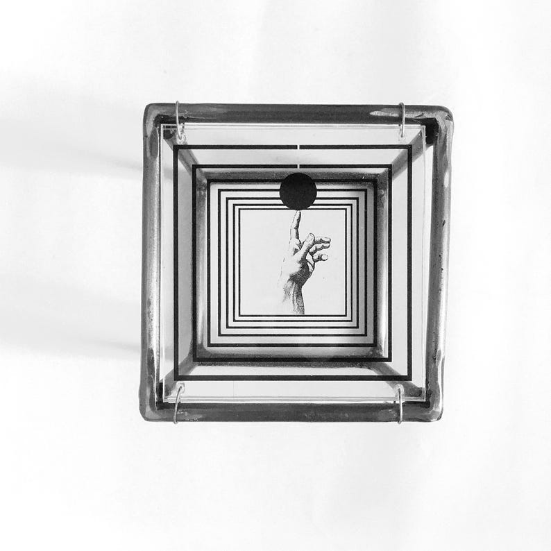 May include: A square glass box with a metal frame featuring a layered design of black concentric squares. Inside, a black circle is positioned above an illustration of a hand with an extended index finger, creating a visual depth effect.