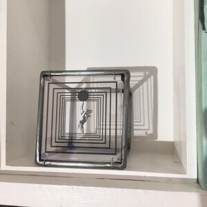 May include: A square metal frame holding a clear acrylic panel with a black graphic print. The print features a series of concentric squares surrounding a central black circle and a detailed illustration of a hand reaching toward the circle.
