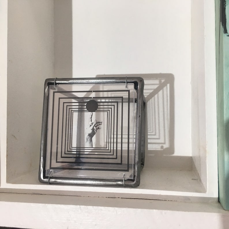 May include: A square metal frame holding a clear acrylic panel with a black graphic print. The print features a series of concentric squares surrounding a central black circle and a detailed illustration of a hand reaching toward the circle.