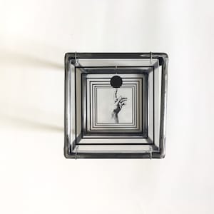 May include: A square glass box constructed with dark metal framing, featuring multiple nested transparent panels printed with black concentric square borders. A central graphic depicts a hand reaching toward a solid black circle.