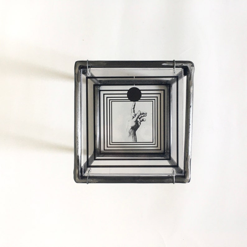 May include: A square glass box constructed with dark metal framing, featuring multiple nested transparent panels printed with black concentric square borders. A central graphic depicts a hand reaching toward a solid black circle.