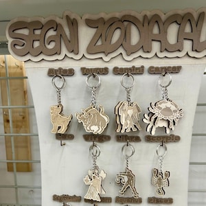 Puede incluir: Un expositor de madera con las palabras "SEGN ZODIACAL" en la parte superior. El expositor presenta 12 signos del zodíaco, cada uno con un llavero. Los signos son Aries, Tauro, Géminis, Cáncer, Leo, Virgo, Libra, Escorpio, Sagitario, Capricornio, Acuario y Piscis.