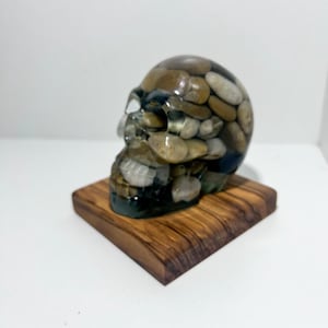 May include: A decorative skull sculpture made of clear resin, filled with small, smooth, tan and brown stones. The skull sits on a small, square, wooden base. The sculpture has a unique, naturalistic appearance.