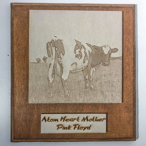 Può includere: Una targa in legno con un'immagine incisa a laser di una mucca in un campo. La targa ha una cornice in legno marrone e uno sfondo bianco. Il testo "Atom Heart Mother Pink Floyd" è inciso sotto l'immagine.