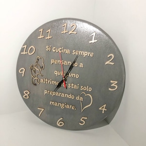 May include: A grey wooden clock with black hands and white numbers. The clock has a quote in Italian that reads "Si cucina sempre pensando a qualcuno altrimenti stai solo preparando da mangiare."
