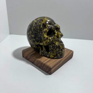 May include: A decorative skull sculpture with a speckled pattern in shades of green, yellow, and black. The skull is displayed on a small, square wooden base. The sculpture has a glossy finish and is set against a white background.