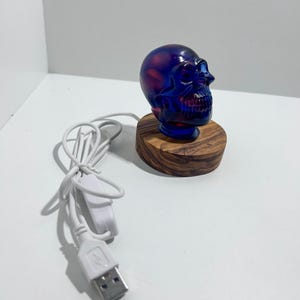 Può includere: Lampada decorativa a forma di teschio, blu e rossa. Il teschio è posto su una piccola base rotonda di legno. Un cavo USB bianco con interruttore è collegato.