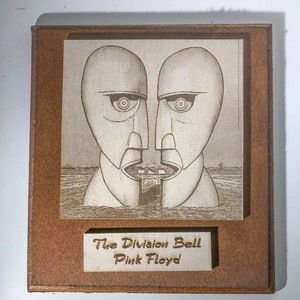 Può includere: Una targa in legno con un'immagine incisa a laser della copertina dell'album "The Division Bell" dei Pink Floyd. La targa ha una cornice in legno marrone e uno sfondo bianco con il titolo dell'album e il nome della band incisi sotto l'immagine.