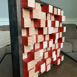 May include: A square wooden wall art piece with a black frame. The wall art is made of small wooden blocks, some painted red and some left natural.
