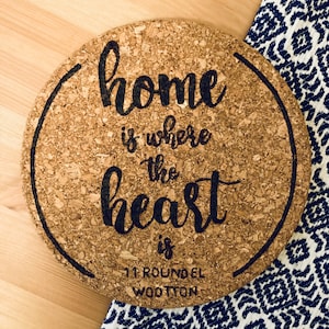 May include: Round cork trivet with the text "home is where the heart is" and the address "11 Roundel Wootton" in black lettering.