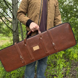 Pode incluir: Uma mala de transporte retangular em pele sintética castanha com acabamento liso, com duas correias verticais com fivelas metálicas, uma pega superior e um pequeno suporte retangular para identificação centrado com rebordo preto.
