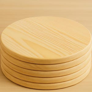 May include: A stack of six round, light-colored wooden coasters. The coasters are made of a light-colored wood with visible grain patterns. The edges are rounded, and the coasters are stacked neatly on a light-colored wooden surface.