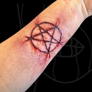 Peut inclure: Un tatouage temporaire représentant un pentagramme noir centré sur la peau, entouré d'une texture de peau rouge et irritée réaliste et d'effets de sang simulés pour créer l'apparence d'une plaie fraîchement incisée sur l'avant-bras.