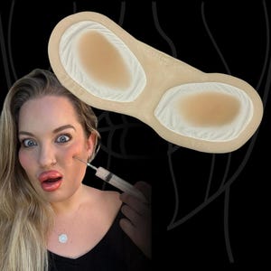 Op de afbeelding: Een beige siliconen prothese met twee ovaalvormige verhoogde delen met getextureerde randen. Het stuk is voorzien van de tekst THE PROSTHETIC GARDEN en LEFT. De prothese is ontworpen voor toepassing op het wanggebied van het gezicht.