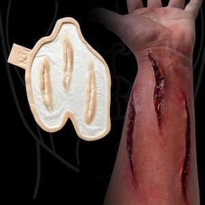 Può includere: Una protesi in silicone con tre lacerazioni parallele su una base color carne, mostrata accanto a un esempio di applicazione su un avambraccio che presenta tre ferite profonde, rosso scuro, piene di sangue simulato.