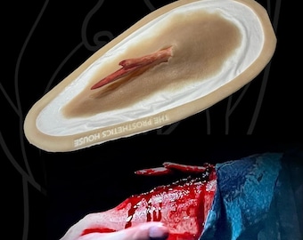 Op de afbeelding: Een siliconen prothese met een realistische huidkleurige ovale basis en een uitstekend splinterachtig object. De tekst THE PROSTHETICS HOUSE staat op de rand gedrukt. Hieronder toont een grafische toepassing een bloedige armwond.