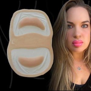 May include: A beige silicone practice mat for lip filler training featuring two moulded lip shapes with a smooth texture. The mat has a central horizontal band and a small letter L embossed on the left side, designed for cosmetic injection technique.
