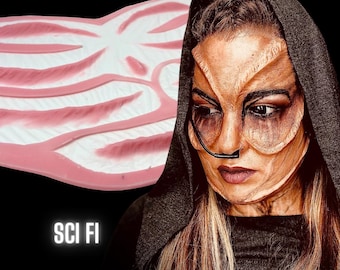 May include: A pink and white silicone mould with a textured design. The mould is shaped like a wing and has the text "SCI FI" printed below it. A person with detailed alien makeup is wearing a black hooded robe.