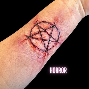 May include: A close-up of a person's arm with a pentagram drawn in black marker. The pentagram is surrounded by red, bloody lines, suggesting a horror or Halloween theme.