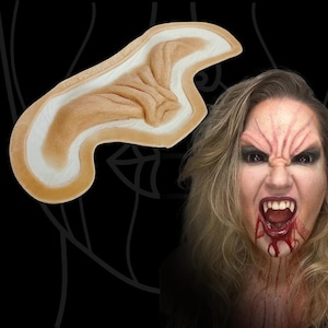 Può includere: Una protesi facciale in silicone beige e testurizzata, caratterizzata da una forma curva e allungata con creste rialzate che imitano la pelle rugosa. L'elemento presenta un bordo liscio e affusolato per una fusione naturale sulla pelle.