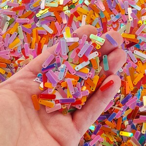 Peut inclure: Gros plan sur une main tenant une multitude de paillettes rectangulaires multicolores. Les paillettes, de couleurs vives, comprennent du rouge, de l'orange, du jaune, du vert, du bleu, du violet et du rose. Chaque paillette a un petit trou.