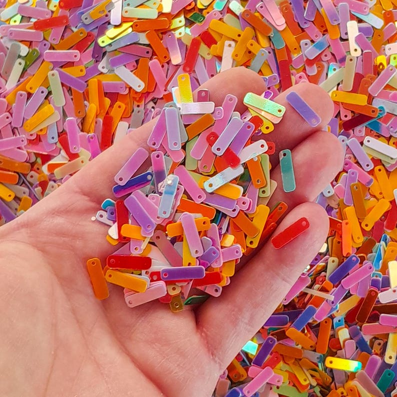 Peut inclure: Gros plan sur une main tenant une multitude de paillettes rectangulaires multicolores. Les paillettes, de couleurs vives, comprennent du rouge, de l'orange, du jaune, du vert, du bleu, du violet et du rose. Chaque paillette a un petit trou.