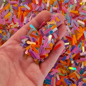 Peut inclure: Gros plan sur une main tenant une poign&eacute;e de paillettes rectangulaires multicolores. Les paillettes sont de diff&eacute;rentes couleurs : orange, rose, violet, jaune et vert. Elles ont un l&eacute;ger &eacute;clat et sont parfaites pour les projets cr&eacute;atifs.