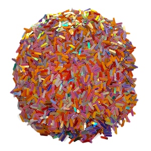 May include: A pile of colorful, iridescent sequins in various shades of orange, pink, purple, and yellow. The sequins are rectangular in shape and scattered across a white background. The sequins have a shiny, reflective surface.