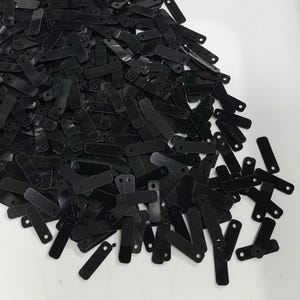 Puede incluir: Un montón grande de lentejuelas rectangulares negras con dos pequeños agujeros en la parte superior. Las lentejuelas están esparcidas sobre una superficie blanca.