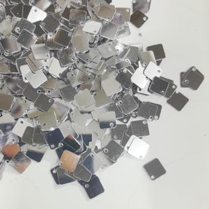 May include: A pile of silver square sequins with a hole in each corner. The sequins are scattered on a white surface.
