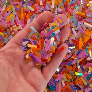 Peut inclure: Une main tenant une collection de paillettes rectangulaires color&eacute;es. Les paillettes sont dans les tons orange, rose, violet, jaune et rouge. Les paillettes ont une finition brillante et iris&eacute;e.