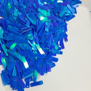 May include: A pile of iridescent blue sequins in the shape of long rectangles. The sequins have a holographic finish and are scattered on a white surface.