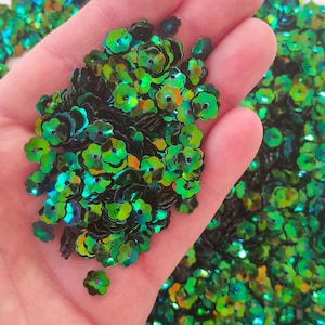 May include: A pile of iridescent green and black flower-shaped sequins. The sequins have a holographic finish and are ideal for crafting and decorating.