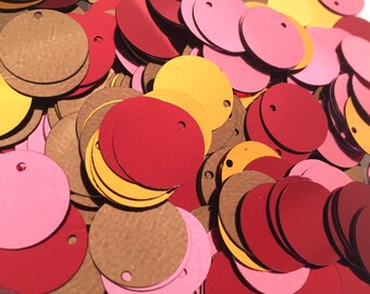 May include: A close-up shot of a pile of colorful, round sequins. The sequins are in shades of red, pink, gold, and brown. Each sequin has a small hole near the edge. The sequins are scattered, creating a textured surface.