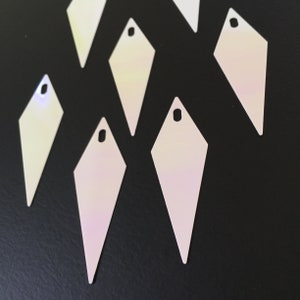 Könnte beinhalten: Eine Gruppe von sieben schimmernden weißen, diamantförmigen Pailletten mit einem Loch oben. Die Pailletten befinden sich auf einem schwarzen Hintergrund.