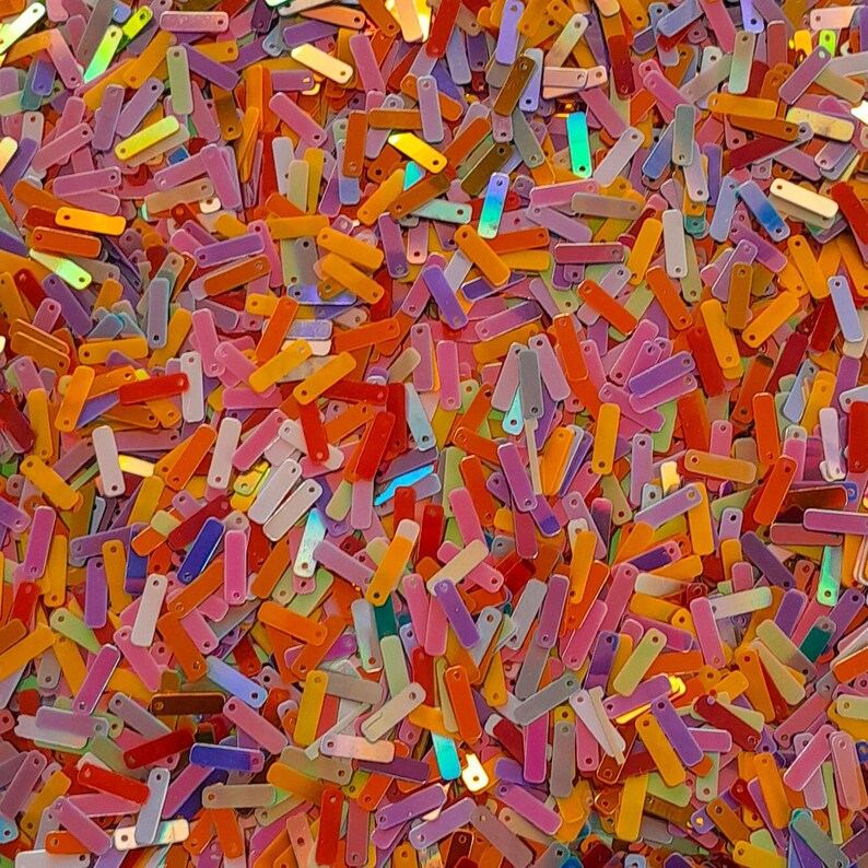 Peut inclure: Gros plan sur une pile de paillettes rectangulaires color&eacute;es. Les paillettes sont dans diverses nuances d'orange, de rose, de jaune, de violet et de vert, cr&eacute;ant une surface vibrante et textur&eacute;e. Les paillettes sont dispers&eacute;es au hasard.